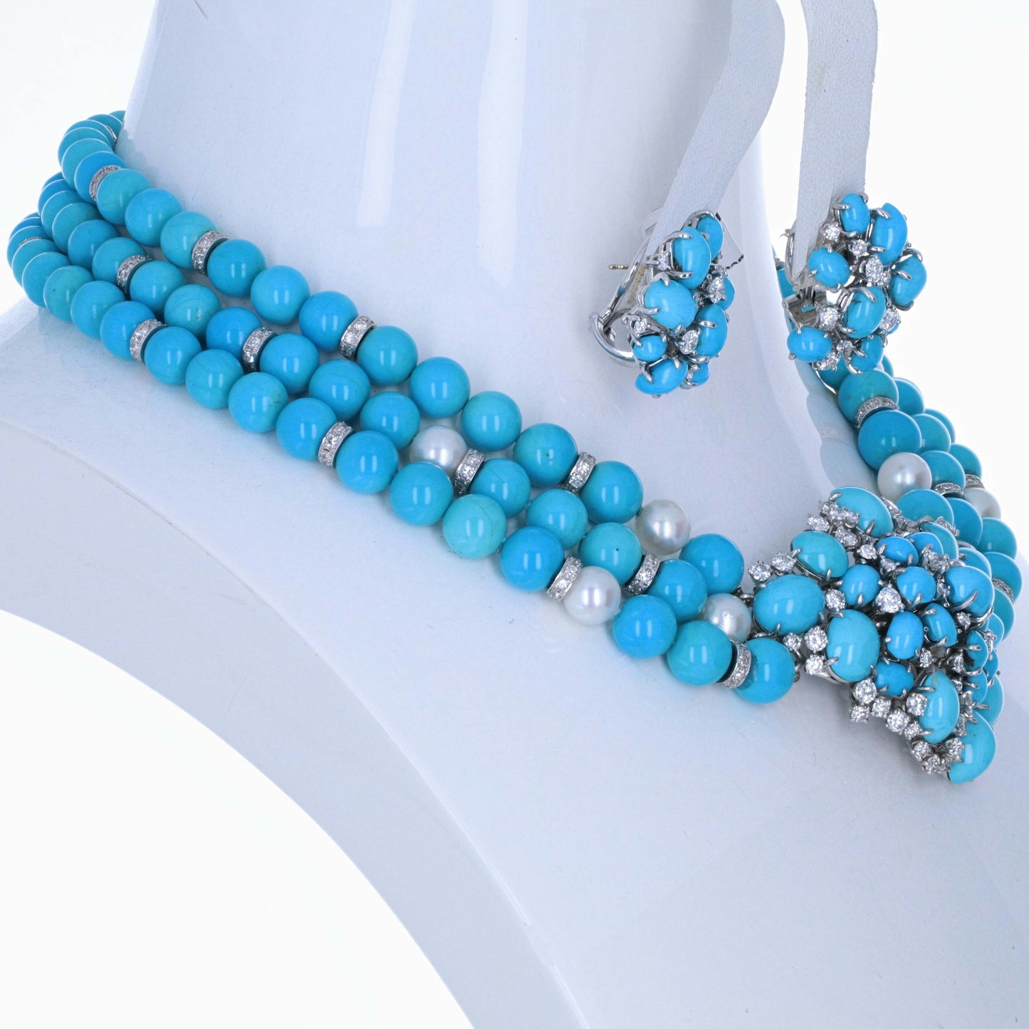meister-swiss-18k-turquoise-diamond-pearl-necklace-earring-suite A turquoise blue beaded jewelry set displayed on a white mannequin bust, including a multi-strand necklace with silver spacer beads and pearl-like accents, matching flower-shaped earrings, and a coordinating bracelet.